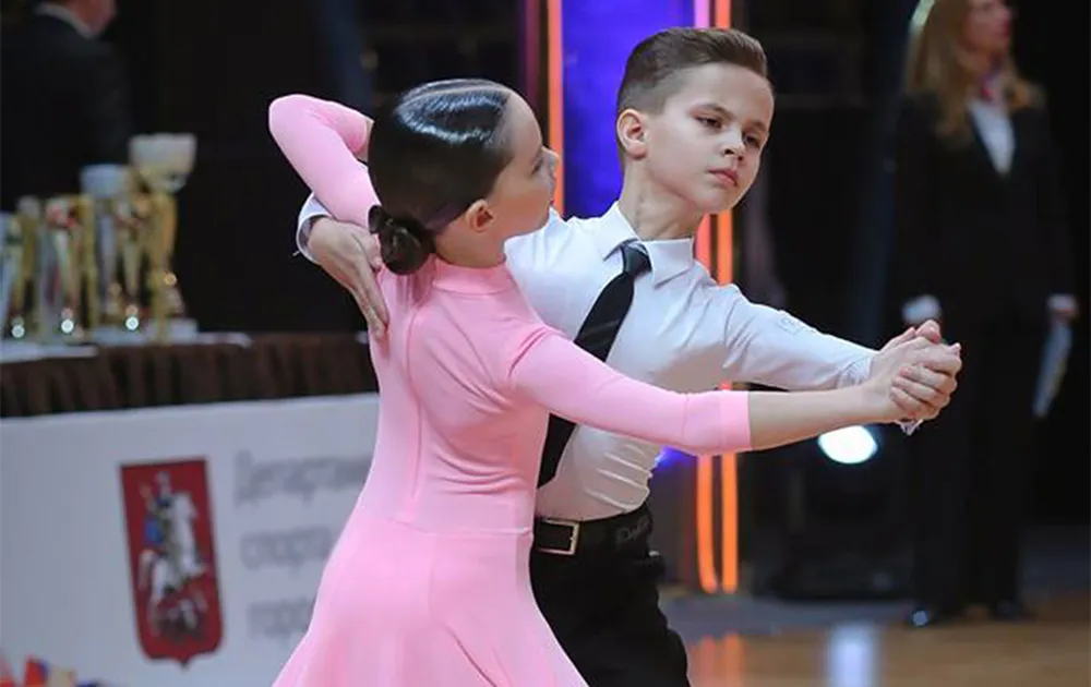 Doi copii dansează dans sportiv la un concurs, ea în rochie roz, el în costum negru